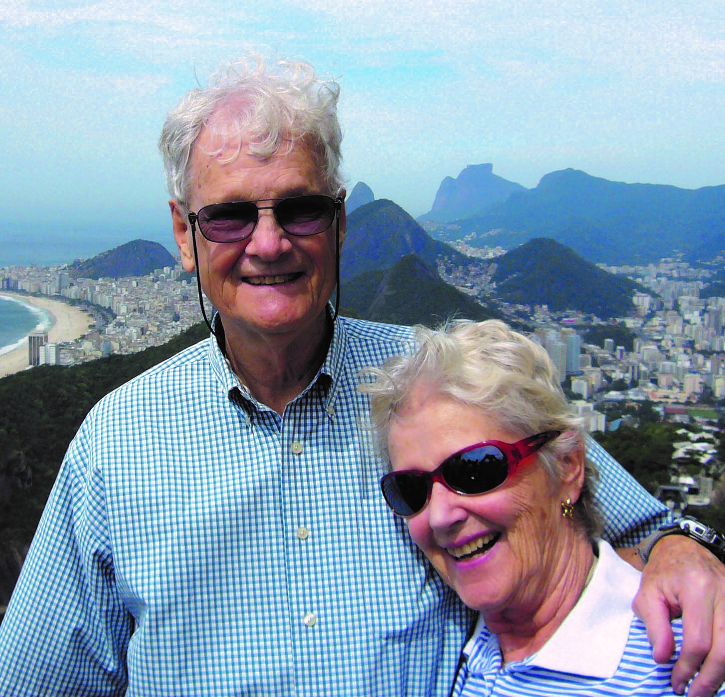 George and Jean Hutchison, an elderly couple in blue and white shirts and sunglasses, smiling, with a mountainous city view behind them